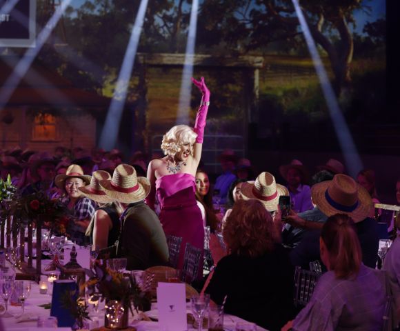 A performer in a vibrant pink dress with matching glove sings amid a lively audience seated at candlelit tables, beneath dramatic stage lighting.