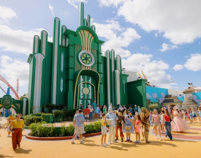 A vibrant theme park scene with visitors enjoying a sunny day. A large, green art deco building is in the background, evoking a whimsical, fantasy atmosphere.