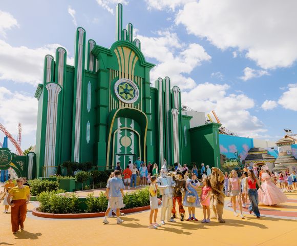 A vibrant theme park scene with visitors enjoying a sunny day. A large, green art deco building is in the background, evoking a whimsical, fantasy atmosphere.