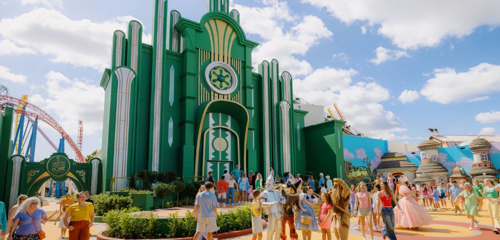 A vibrant theme park scene with visitors enjoying a sunny day. A large, green art deco building is in the background, evoking a whimsical, fantasy atmosphere.