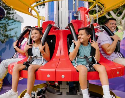 Children and adults smiling on a spinning amusement park ride with red seats and safety harnesses. The background shows colorful structures.