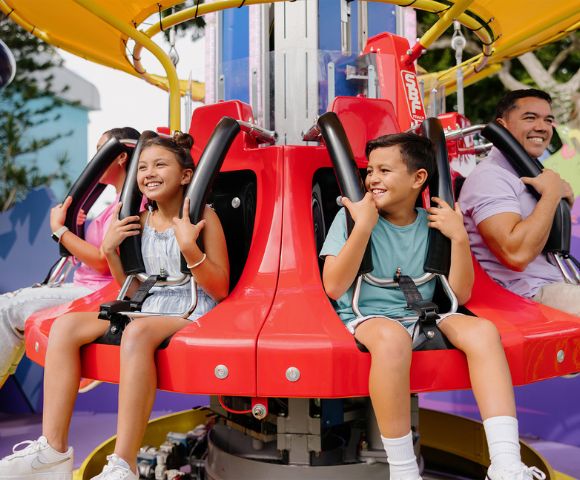 Children and adults smiling on a spinning amusement park ride with red seats and safety harnesses. The background shows colorful structures.