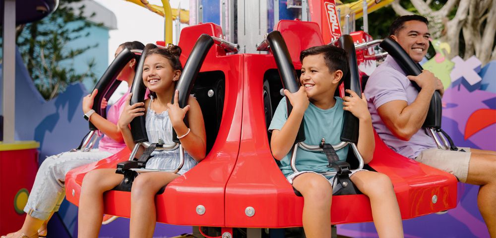 Children and adults smiling on a spinning amusement park ride with red seats and safety harnesses. The background shows colorful structures.