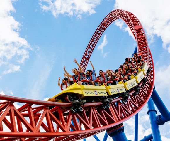 A vibrant red and yellow roller coaster soars through a loop against a bright blue sky with scattered clouds. Riders, arms raised, exude excitement and thrill.