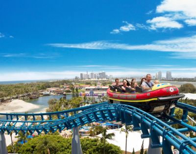 A colorful roller coaster with a group of excited riders ascends a track above a scenic park. The sky is clear blue with city skyline in the distance.