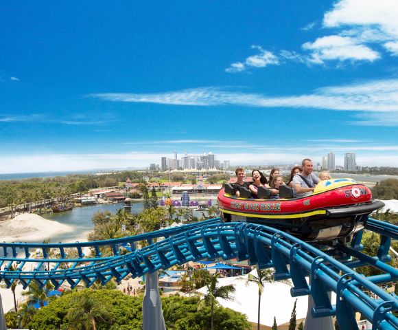 A colorful roller coaster with a group of excited riders ascends a track above a scenic park. The sky is clear blue with city skyline in the distance.