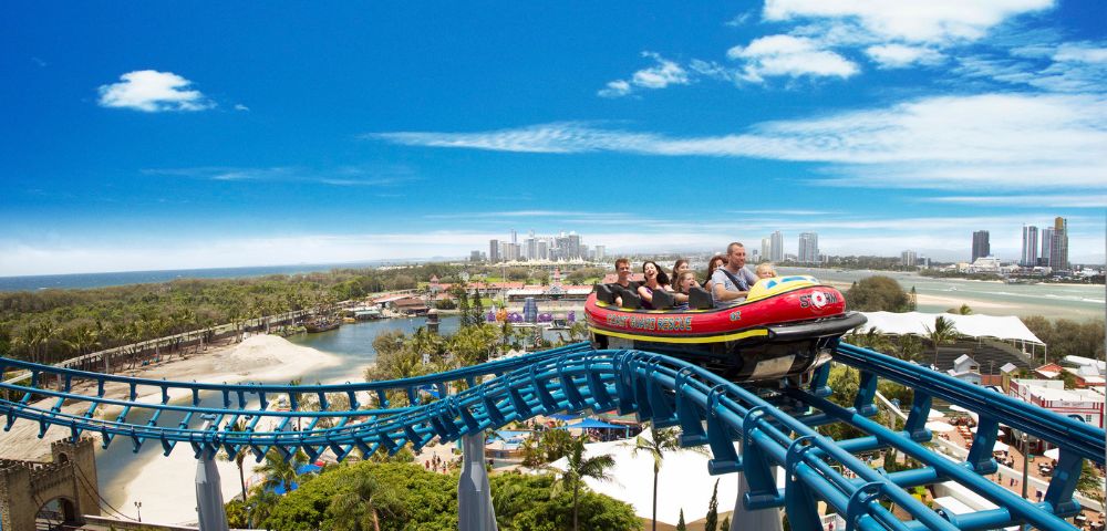 A colorful roller coaster with a group of excited riders ascends a track above a scenic park. The sky is clear blue with city skyline in the distance.