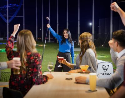 A woman triumphantly raises a golf club at a Topgolf venue, surrounded by friends cheering with drinks, conveying joy and celebration.
