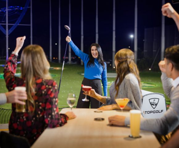 A woman triumphantly raises a golf club at a Topgolf venue, surrounded by friends cheering with drinks, conveying joy and celebration.