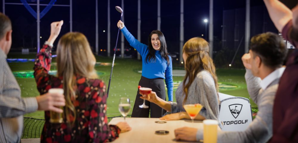 A woman triumphantly raises a golf club at a Topgolf venue, surrounded by friends cheering with drinks, conveying joy and celebration.