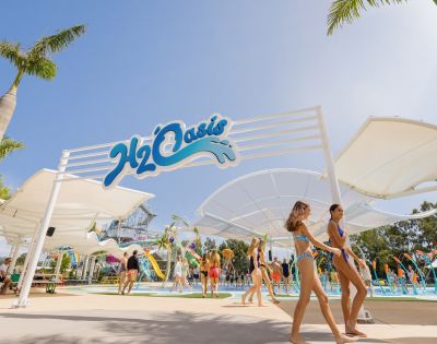 People in swimsuits enjoy a sunny day at a water park named H2Oasis. The scene includes slides and pools, evoking a lively and fun atmosphere.