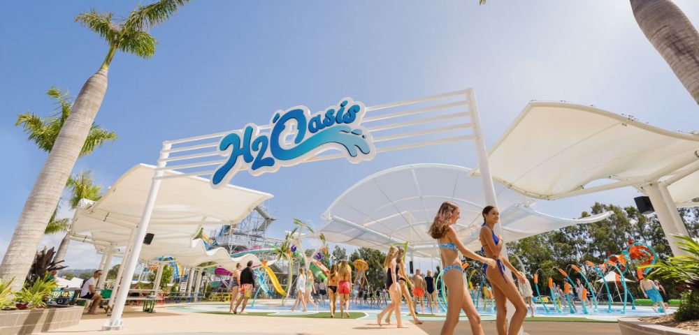 People in swimsuits enjoy a sunny day at a water park named H2Oasis. The scene includes slides and pools, evoking a lively and fun atmosphere.