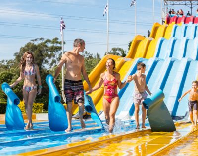 A family enjoying a sunny day at a water park, carrying blue mats. They smile as they walk away from a large, colorful slide, conveying fun and excitement.