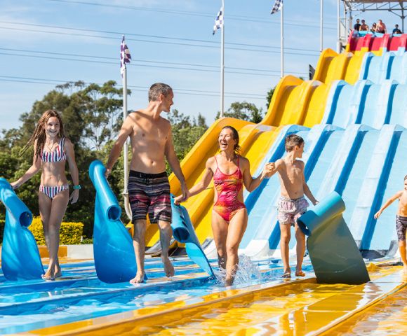 A family enjoying a sunny day at a water park, carrying blue mats. They smile as they walk away from a large, colorful slide, conveying fun and excitement.