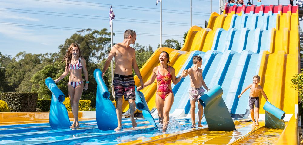 A family enjoying a sunny day at a water park, carrying blue mats. They smile as they walk away from a large, colorful slide, conveying fun and excitement.