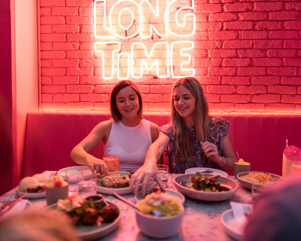 Two women sitting at a table with various dishes and drinks, under a neon sign that reads 