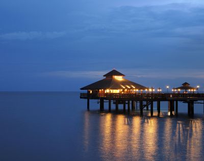 Wooden pavilion on stilts over calm sea at dusk, warmly lit with glowing lights reflecting on the water. The sky is twilight blue, evoking a serene mood.