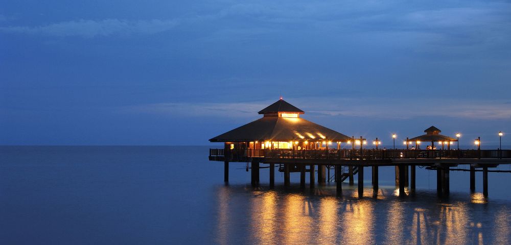 Wooden pavilion on stilts over calm sea at dusk, warmly lit with glowing lights reflecting on the water. The sky is twilight blue, evoking a serene mood.