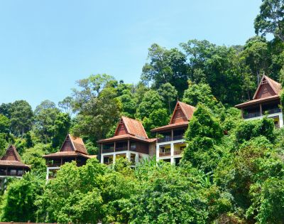 Row of traditional wooden villas with steep roofs set amidst lush green trees on a hillside under a clear blue sky, conveying a serene, tropical retreat.