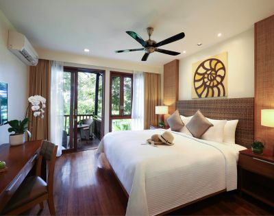 Bright hotel room with a large bed, decorative pillows, and wall art above. Balcony doors open to a lush garden. Warm lighting and a ceiling fan create a cozy atmosphere.