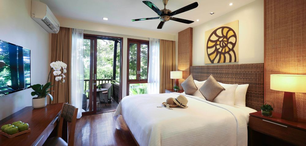 Bright hotel room with a large bed, decorative pillows, and wall art above. Balcony doors open to a lush garden. Warm lighting and a ceiling fan create a cozy atmosphere.