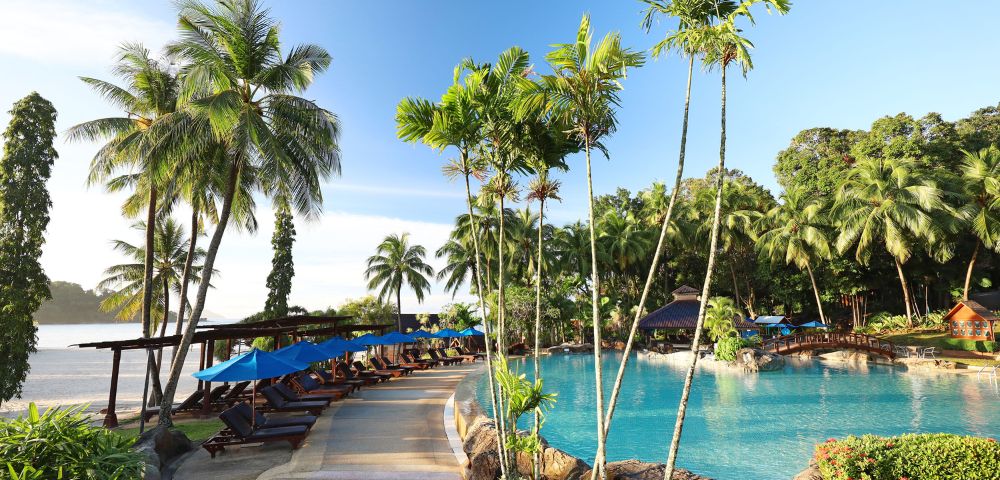Tropical resort poolside with tall palm trees, clear blue water, and sun loungers under blue umbrellas. The setting conveys relaxation and tranquility.
