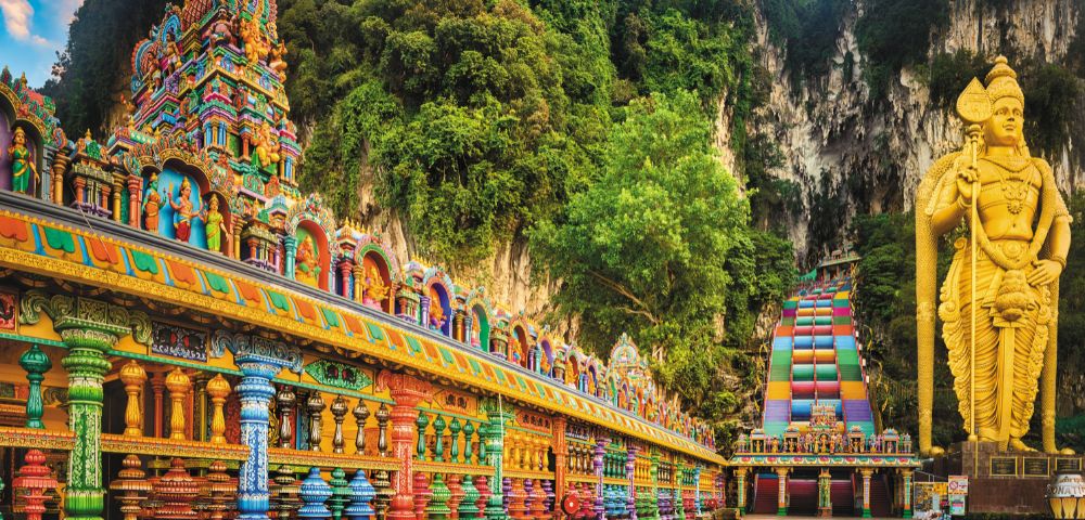 A vibrant Hindu temple with intricate, colorful carvings leads to a towering, gold statue amidst lush greenery and rocky cliffs, conveying awe and spirituality.