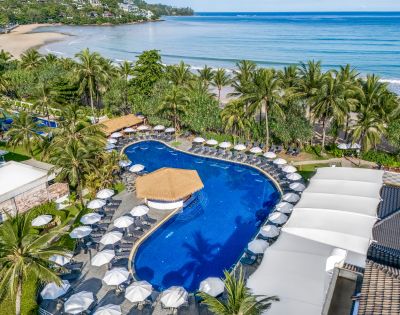Aerial view of a tropical beach resort with a large, curved swimming pool surrounded by white umbrellas and palm trees. Ocean waves in the background.