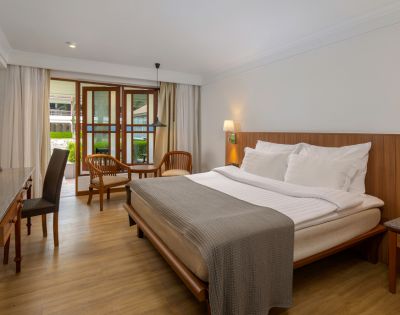 A cozy hotel room with a large bed, soft white bedding, and a gray throw. Wooden furnishings include a desk, chairs, and a small nightstand. Natural light filters through sheer curtains, creating a warm, inviting atmosphere.