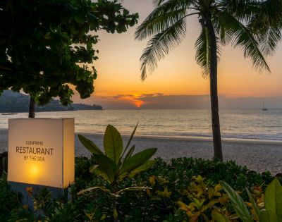 Sunset view at a beachside restaurant, framed by lush greenery and palm trees, with a glowing sky, calm ocean, and sailboat in the distance.