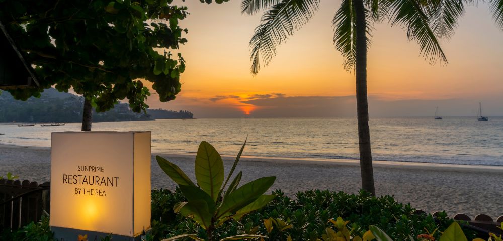 Sunset view at a beachside restaurant, framed by lush greenery and palm trees, with a glowing sky, calm ocean, and sailboat in the distance.