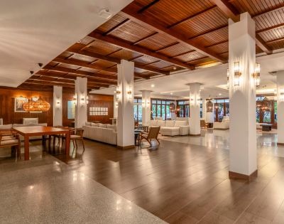 Spacious hotel lobby with warm lighting, wooden ceiling, and white pillars. Cozy seating areas with white couches create an inviting and elegant atmosphere.