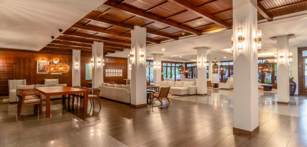 Spacious hotel lobby with warm lighting, wooden ceiling, and white pillars. Cozy seating areas with white couches create an inviting and elegant atmosphere.