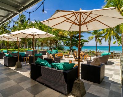 Tropical beachfront patio with wicker furniture, blue cushions, and large white umbrellas. Palm trees and ocean in the background under a clear blue sky.
