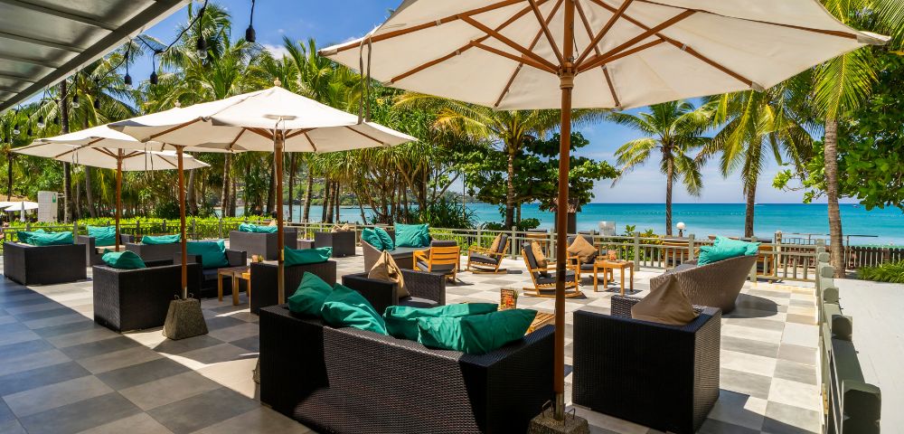 Tropical beachfront patio with wicker furniture, blue cushions, and large white umbrellas. Palm trees and ocean in the background under a clear blue sky.