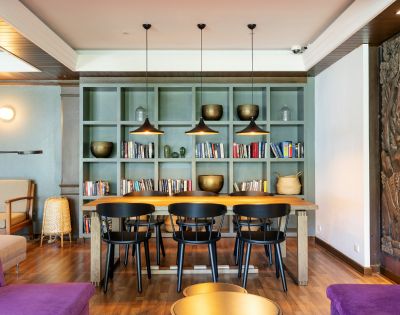 Cozy room with a wooden table, five black chairs, and an olive bookshelf filled with books and decorative objects. Warm, modern ambiance with pendant lights.