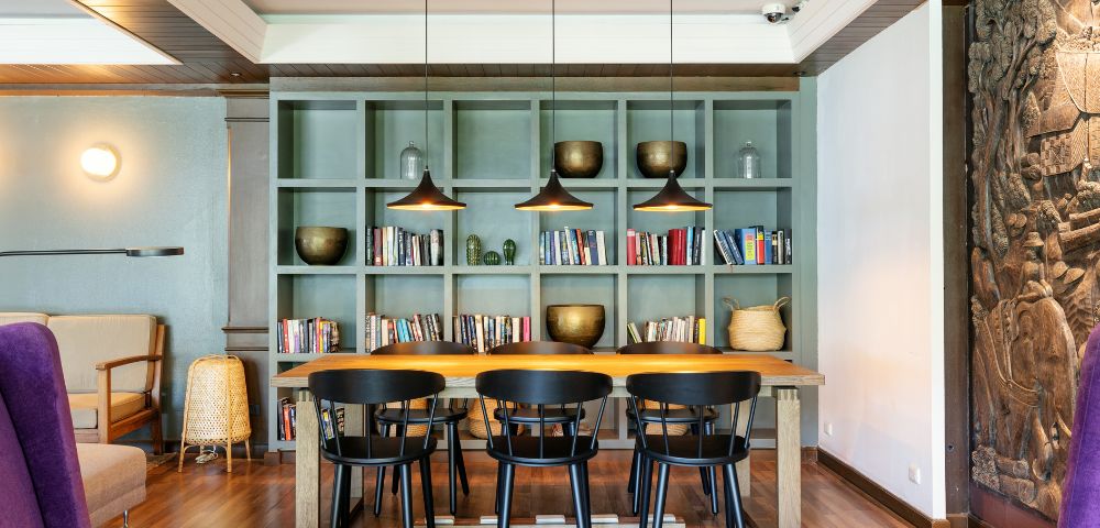 Cozy room with a wooden table, five black chairs, and an olive bookshelf filled with books and decorative objects. Warm, modern ambiance with pendant lights.