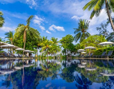 Tropical pool scene with clear blue water reflecting palm trees and umbrellas under a vibrant sky. Relaxing, serene atmosphere.