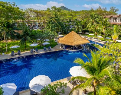 A serene tropical resort features a large azure pool with a central straw-roofed bar, surrounded by lush greenery, lounge chairs, and white umbrellas.