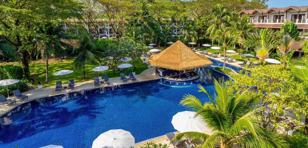 A serene tropical resort features a large azure pool with a central straw-roofed bar, surrounded by lush greenery, lounge chairs, and white umbrellas.