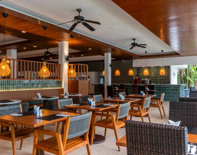 A modern restaurant with warm lighting, wooden ceiling, wicker chairs, and wooden tables set for dining. The atmosphere is inviting and cozy.