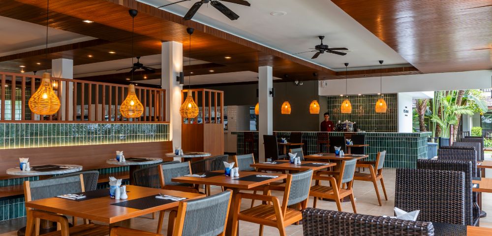 A modern restaurant with warm lighting, wooden ceiling, wicker chairs, and wooden tables set for dining. The atmosphere is inviting and cozy.