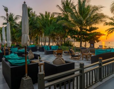 Outdoor patio with plush seating and umbrellas, set against a backdrop of palm trees and a vibrant sunset over the ocean, creating a relaxing atmosphere.