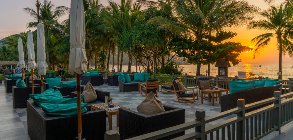 Outdoor patio with plush seating and umbrellas, set against a backdrop of palm trees and a vibrant sunset over the ocean, creating a relaxing atmosphere.