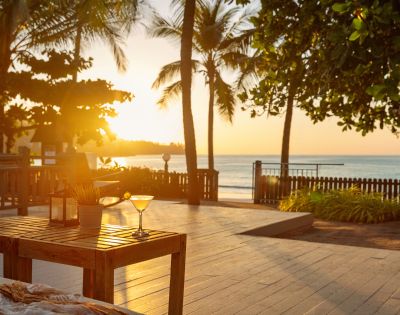 Sunset over a serene beach with palm trees. A wooden table in the foreground holds a drink, creating a tranquil, tropical atmosphere.