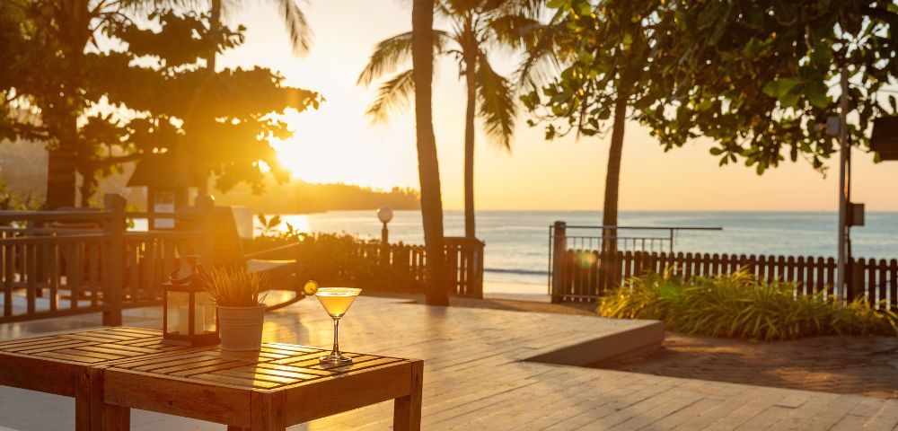 Sunset over a serene beach with palm trees. A wooden table in the foreground holds a drink, creating a tranquil, tropical atmosphere.