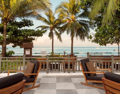 Cozy beachside scene with wooden rocking chairs on a patio, overlooking palm trees and a serene ocean view, evoking a relaxing tropical vibe.