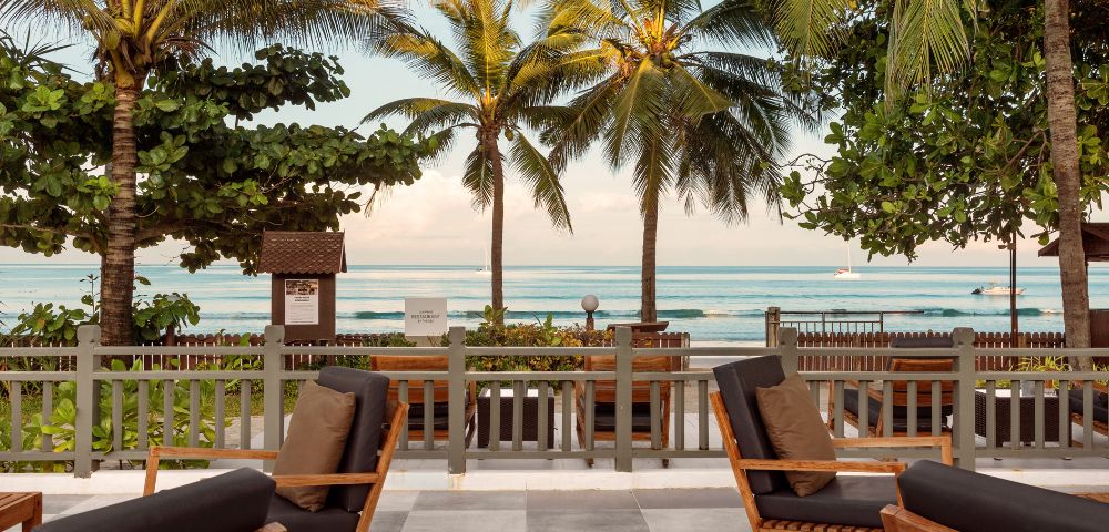 Cozy beachside scene with wooden rocking chairs on a patio, overlooking palm trees and a serene ocean view, evoking a relaxing tropical vibe.