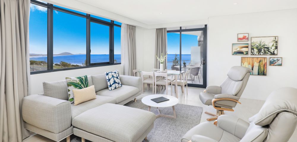Modern, bright living room with large windows showcasing ocean views. Features a gray sectional sofa, cozy armchairs, and a dining table, creating a serene atmosphere.