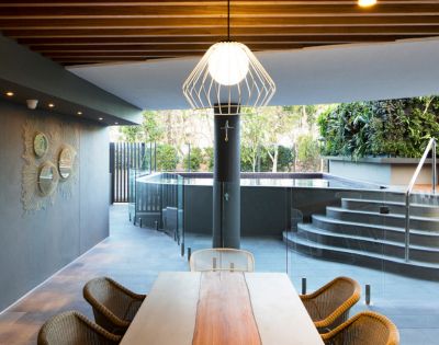 Stylish outdoor dining area with wooden table and wicker chairs. A modern pendant light hangs above, leading to a sleek pool with steps and lush greenery.